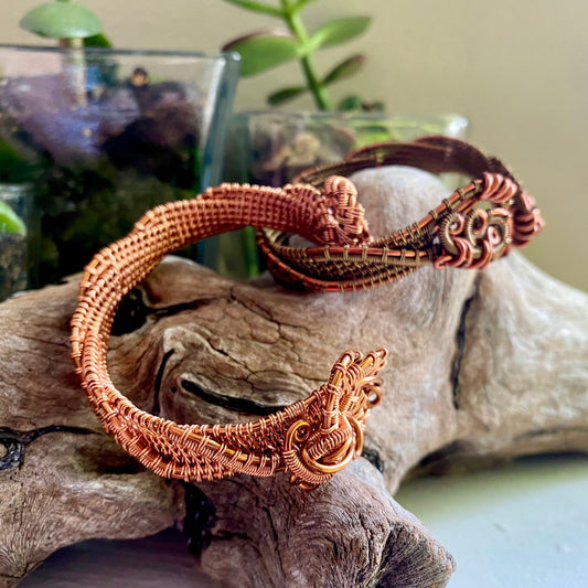 Two twisting cuff bracelets resting on driftwood with plants in background. Wire wrapped cuff are in copper and a mix of copper and antique bronze.